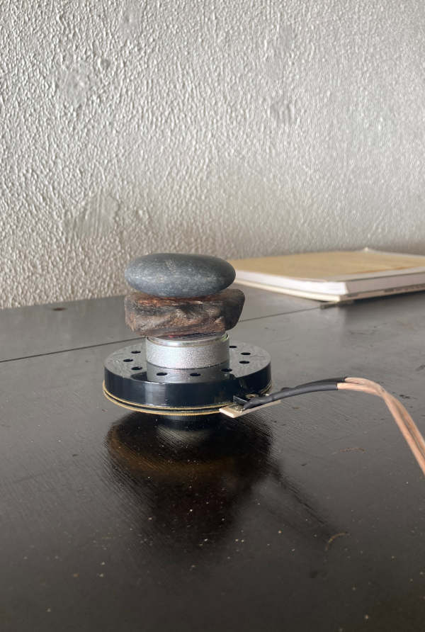 A transducer (exciter) with stones piled on top of it sitting on the top of an upright piano against a white wall.